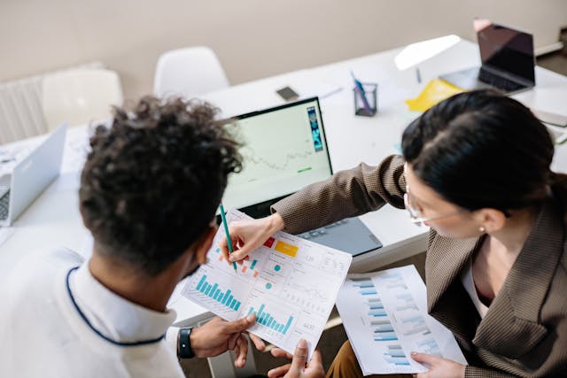 two people in an office setting looking at charts both in a hardcopy version and on the screen of a laptop